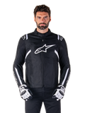Alpinestars T-SPS V2 Waterproof Jacket en noir et blanc, vue de face sur modèle, veste de moto en textile imperméable avec des bandes blanches sur les épaules et les bras, grand logo Alpinestars blanc sur la poitrine, portée avec des gants de moto protecteurs, silhouette élégante d'inspiration sportive