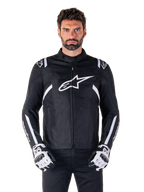 Alpinestars T-SPS V2 Waterproof Jacket en noir et blanc, vue de face sur modèle, veste de moto en textile imperméable avec des bandes blanches sur les épaules et les bras, grand logo Alpinestars blanc sur la poitrine, portée avec des gants de moto protecteurs, silhouette élégante d'inspiration sportive