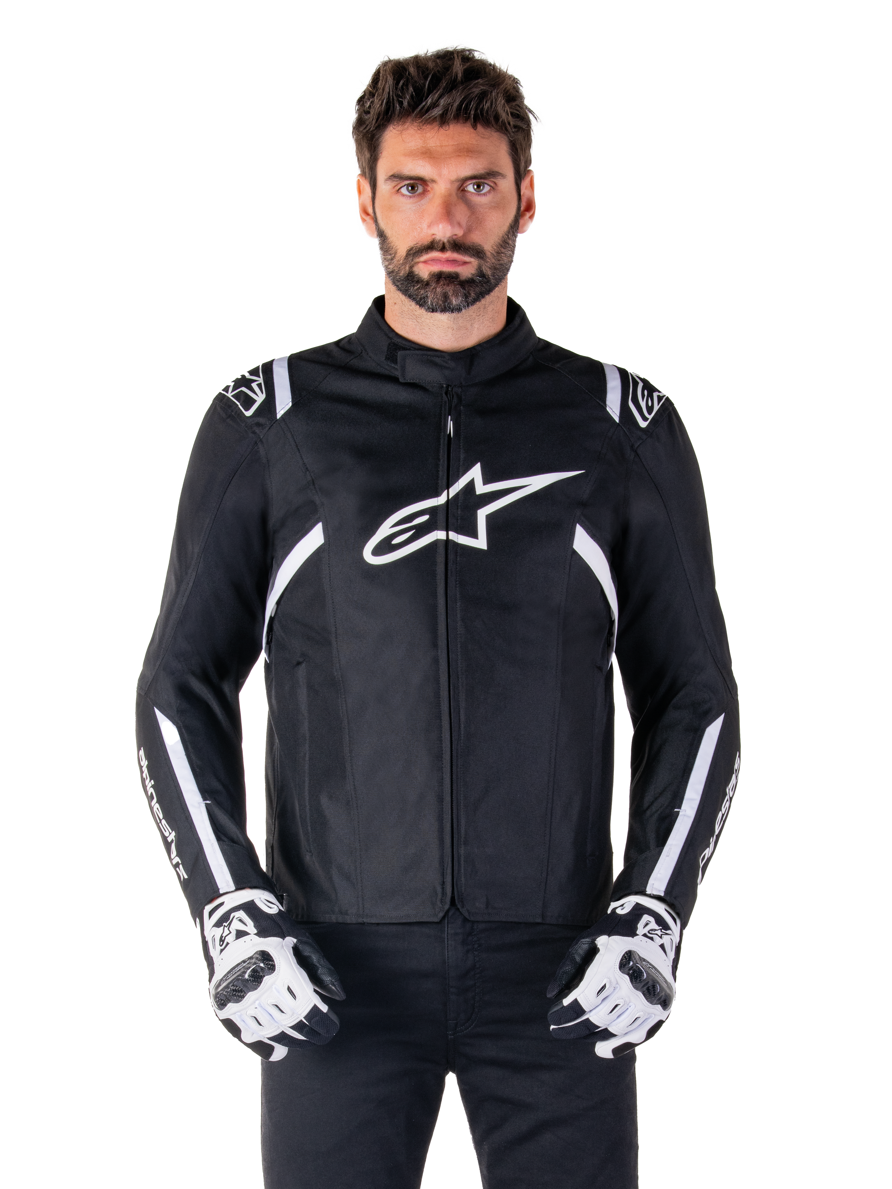 Alpinestars T-SPS V2 Waterproof Jacket en noir et blanc, vue de face sur modèle, veste de moto en textile imperméable avec des bandes blanches sur les épaules et les bras, grand logo Alpinestars blanc sur la poitrine, portée avec des gants de moto protecteurs, silhouette élégante d'inspiration sportive
