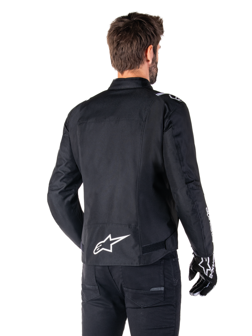 Veste imperméable Alpinestars T-SPS V2, noir et blanc, vue arrière d'un motard portant la veste, logo Alpinestars blanc dans le bas du dos, construction textile à coupe sport, portée avec un pantalon et des gants de moto noirs