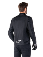Veste imperméable Alpinestars T-SPS V2, noir et blanc, vue arrière d'un motard portant la veste, logo Alpinestars blanc dans le bas du dos, construction textile à coupe sport, portée avec un pantalon et des gants de moto noirs