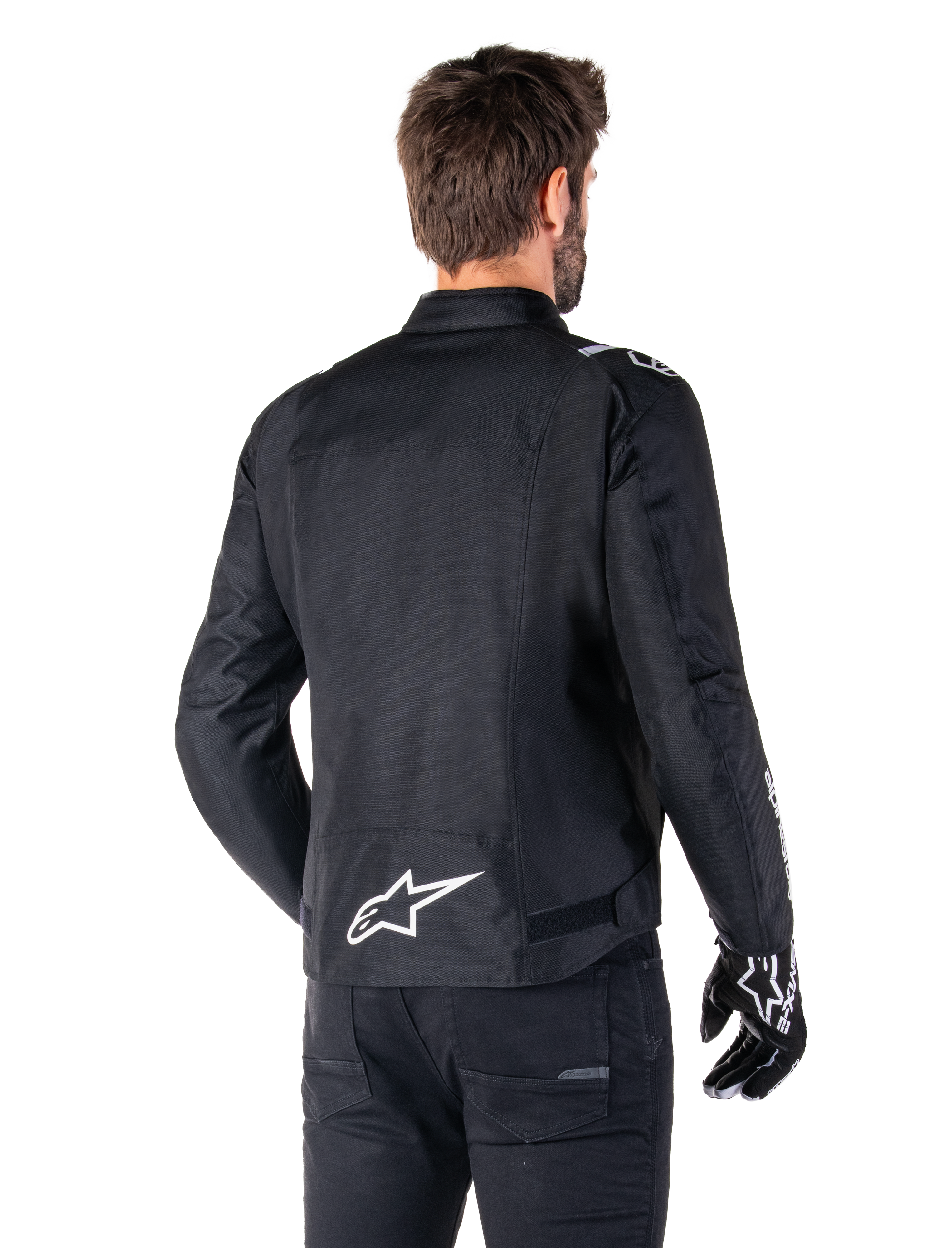 Veste imperméable Alpinestars T-SPS V2, noir et blanc, vue arrière d'un motard portant la veste, logo Alpinestars blanc dans le bas du dos, construction textile à coupe sport, portée avec un pantalon et des gants de moto noirs