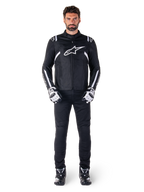 Veste imperméable Alpinestars T-SPS V2, noir et blanc, portée par un mannequin homme avec un pantalon noir et des gants assortis, avec le logo Alpinestars blanc sur la poitrine et des détails sur les manches, silhouette à coupe sport pour la conduite de moto par tous les temps