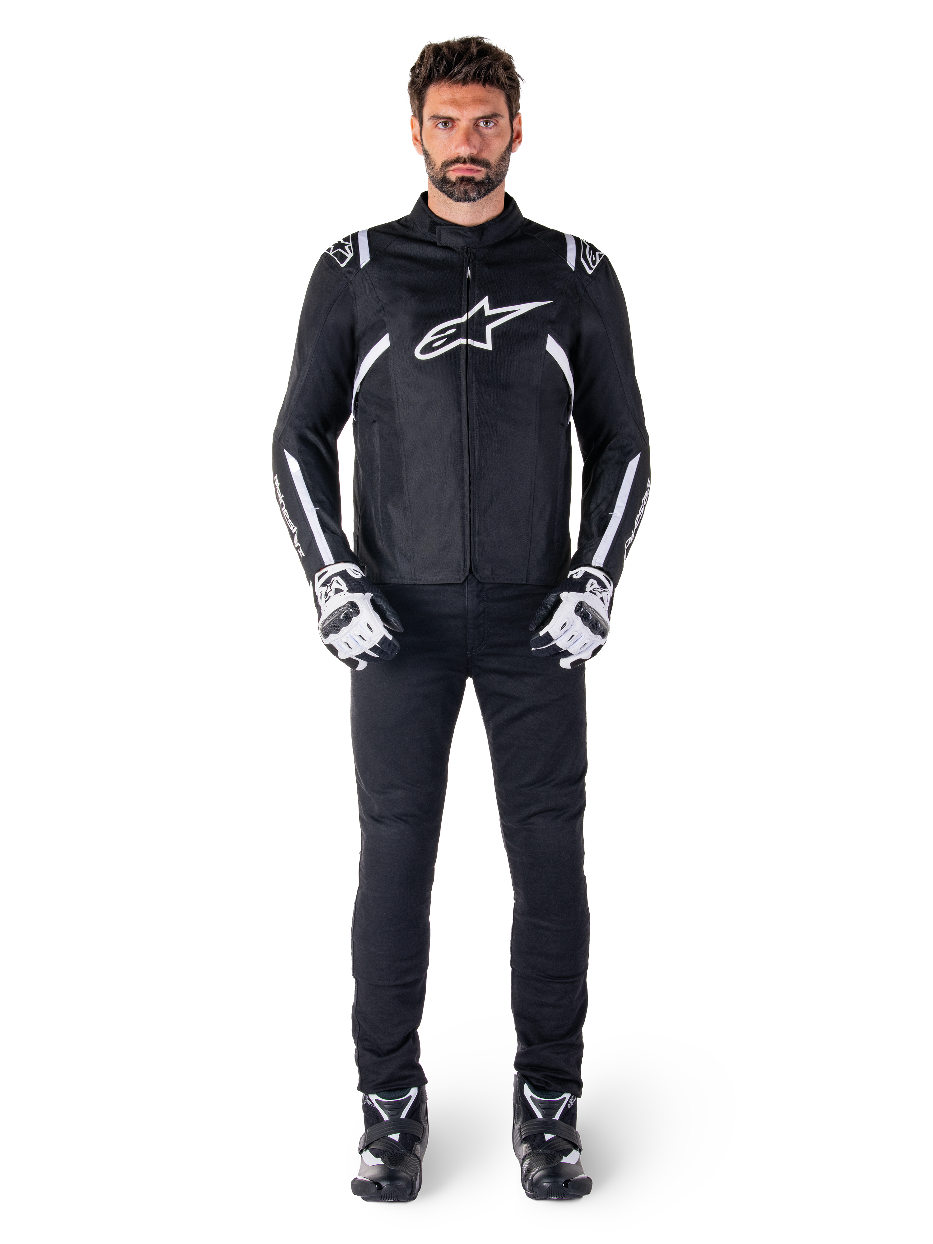 Veste imperméable Alpinestars T-SPS V2, noir et blanc, portée par un mannequin homme avec un pantalon noir et des gants assortis, avec le logo Alpinestars blanc sur la poitrine et des détails sur les manches, silhouette à coupe sport pour la conduite de moto par tous les temps