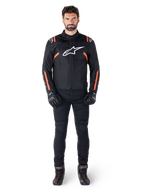 Modèle masculin portant la veste imperméable Alpinestars T-SPS V2 en noir, blanc et rouge fluo, associée à un pantalon de moto et des gants noirs, avec un logo Alpinestars blanc sur la poitrine et des accents rouges sur les manches.