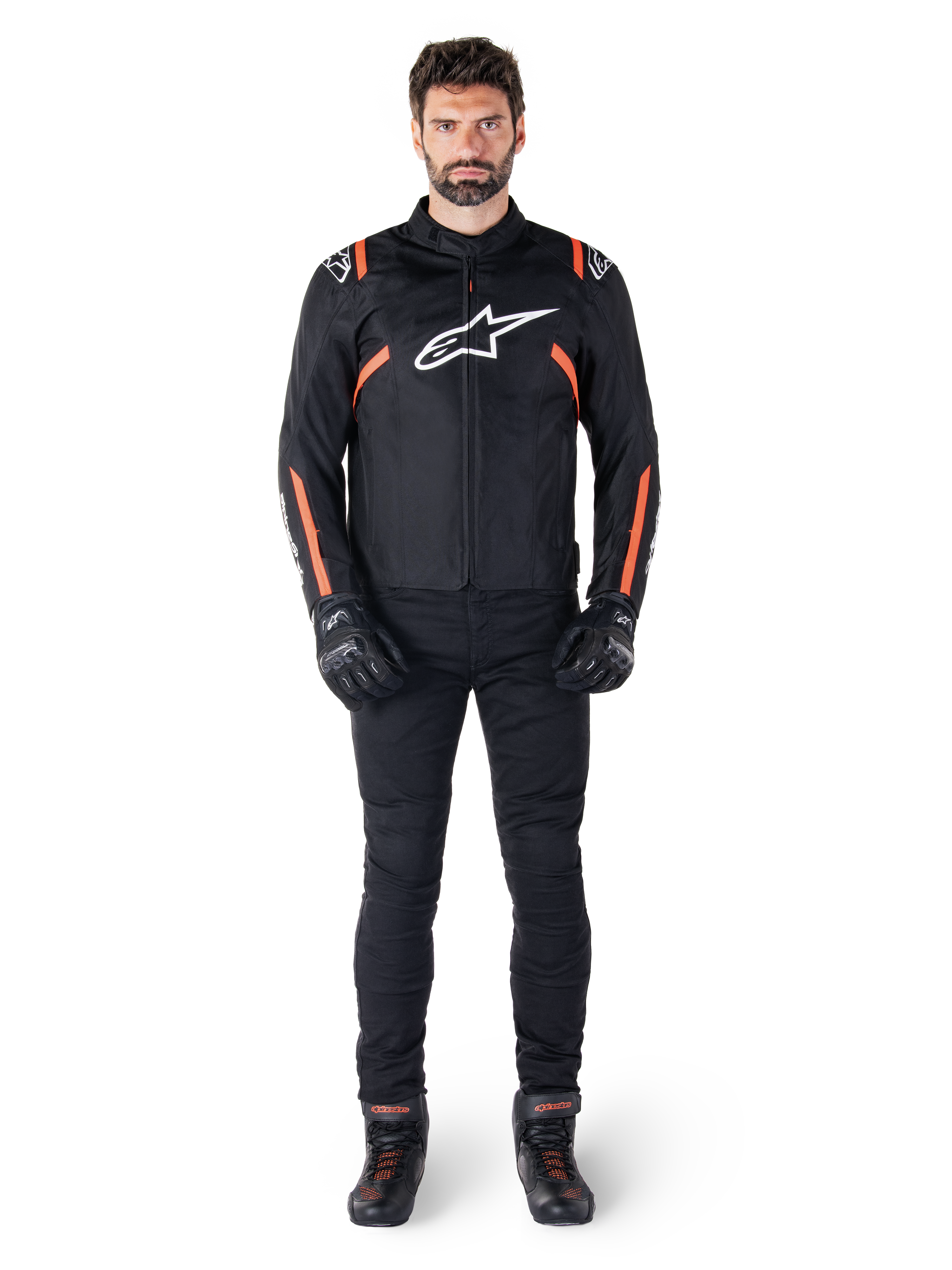 Modèle masculin portant la veste imperméable Alpinestars T-SPS V2 en noir, blanc et rouge fluo, associée à un pantalon de moto et des gants noirs, avec un logo Alpinestars blanc sur la poitrine et des accents rouges sur les manches.