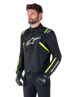 Male model wearing the Alpinestars T-SPS V2 Waterproof Jacket, Black with White and Yellow Fluo accents, featuring a white Astars logo on the chest and fluorescent yellow stripes on the shoulders and sleeves, paired with black pants and riding gloves, sport-cut textile motorcycle jacket.