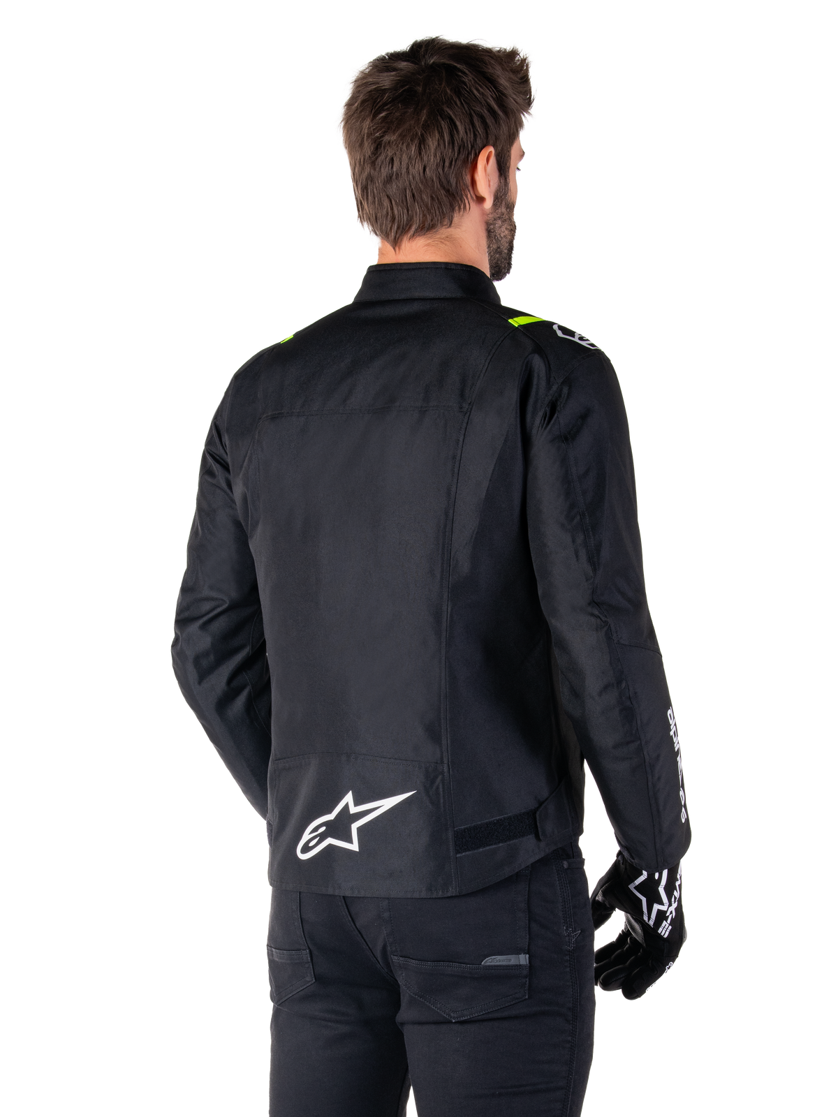 Veste imperméable Alpinestars T-SPS V2, Noir Blanc Jaune Fluo, vue arrière d'un modèle masculin portant la veste de moto en textile, avec un logo blanc en bas du dos et des accents jaune fluo sur les épaules, associée à un pantalon et des gants noirs.