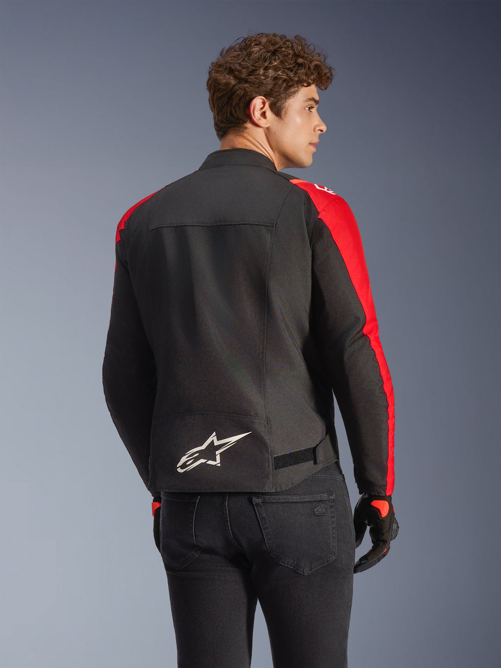 Veste imperméable Alpinestars T-SPS V2 en noir et rouge, vue de dos sur un mannequin, présentant un logo Alpinestars blanc dans le bas du dos, des empiècements de manches rouge vif et une silhouette à coupe sport, portée avec des gants noirs et un jean.