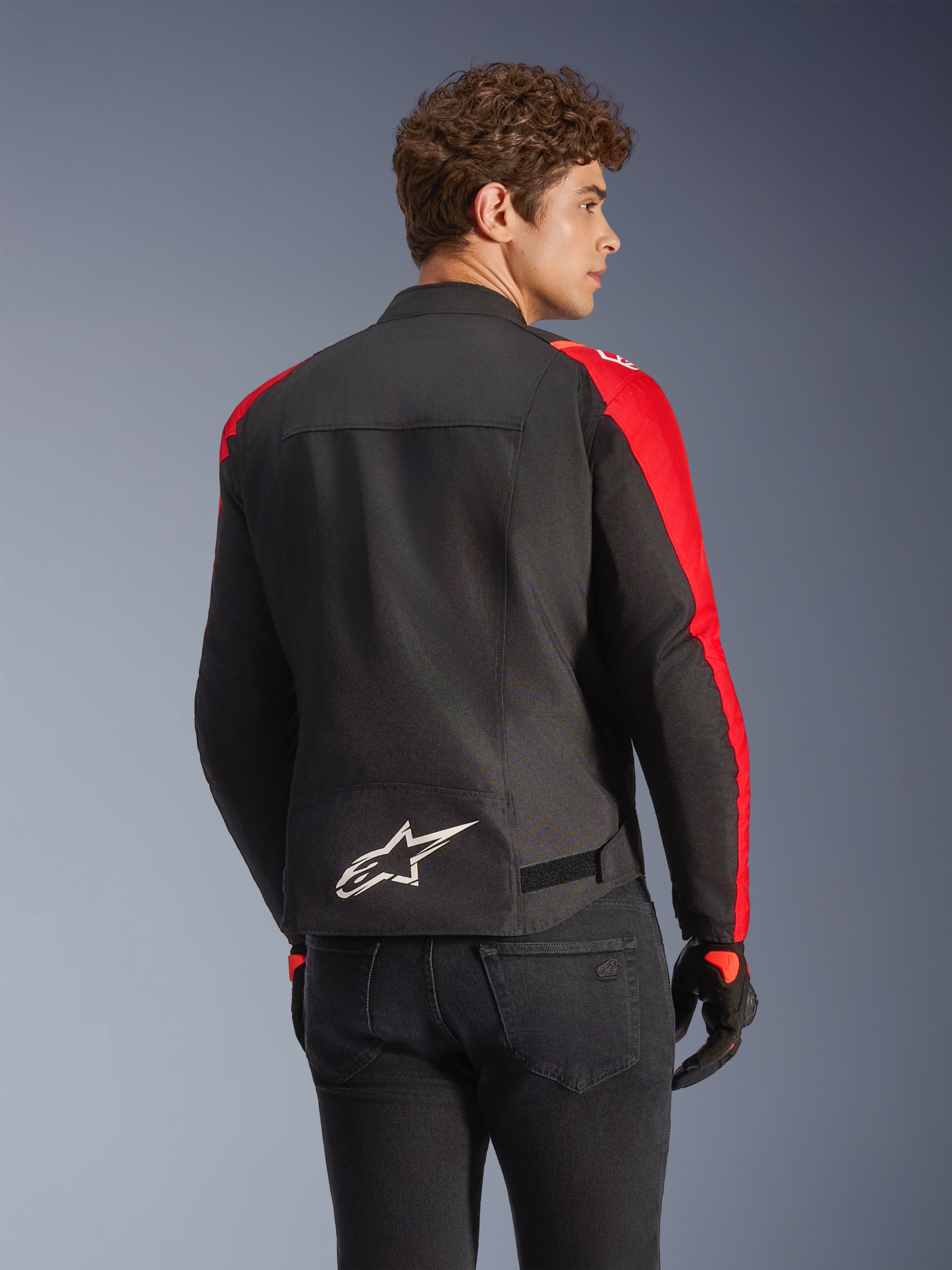 Veste imperméable Alpinestars T-SPS V2 en noir et rouge, vue de dos sur un mannequin, présentant un logo Alpinestars blanc dans le bas du dos, des empiècements de manches rouge vif et une silhouette à coupe sport, portée avec des gants noirs et un jean.