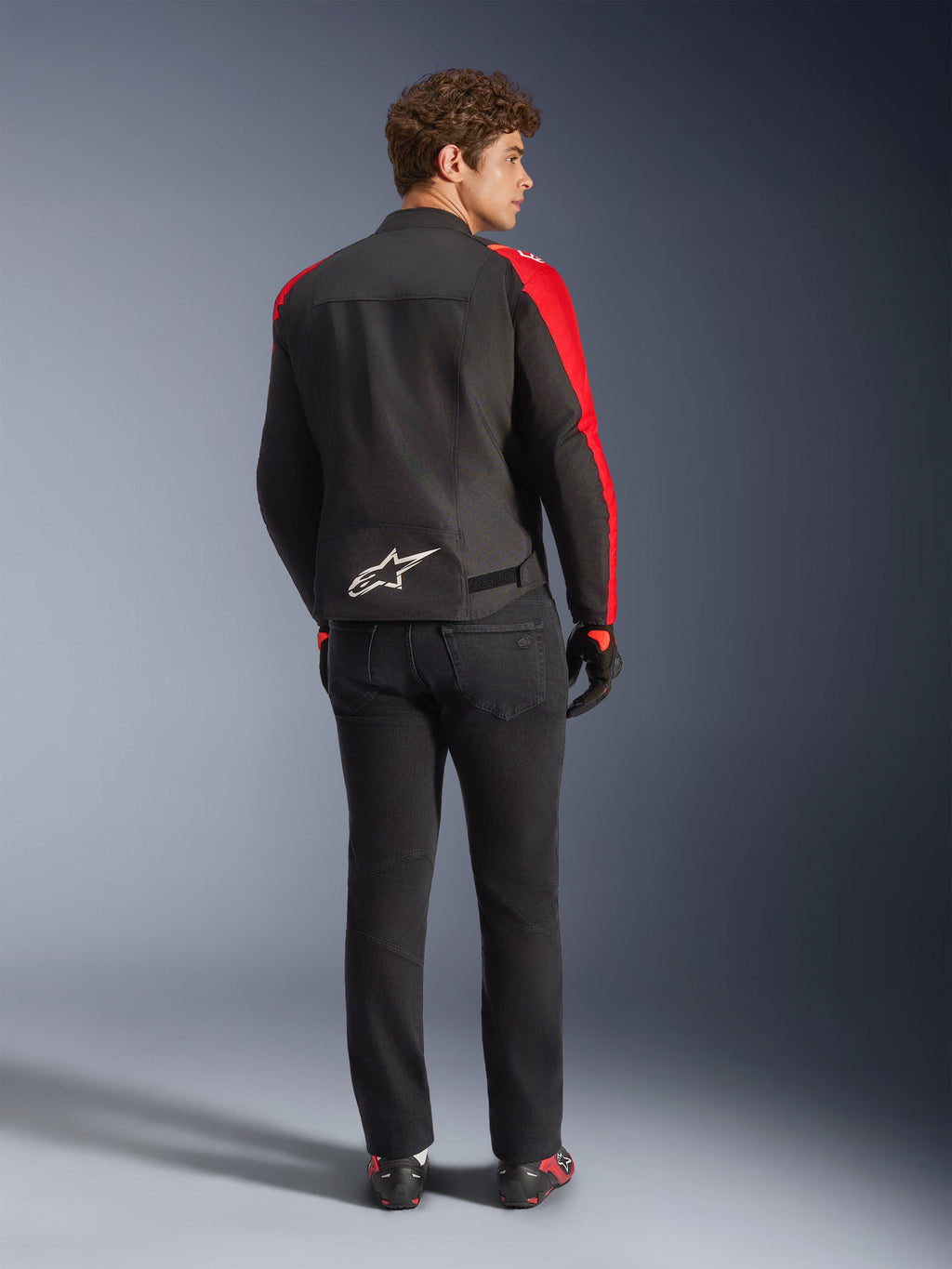 Veste imperméable Alpinestars T-SPS V2, noir, rouge et rouge fluo, vue de dos sur un modèle masculin, logo Alpinestars blanc en bas du dos, portée avec un pantalon de moto noir et des gants, veste de moto en textile imperméable au style sportif