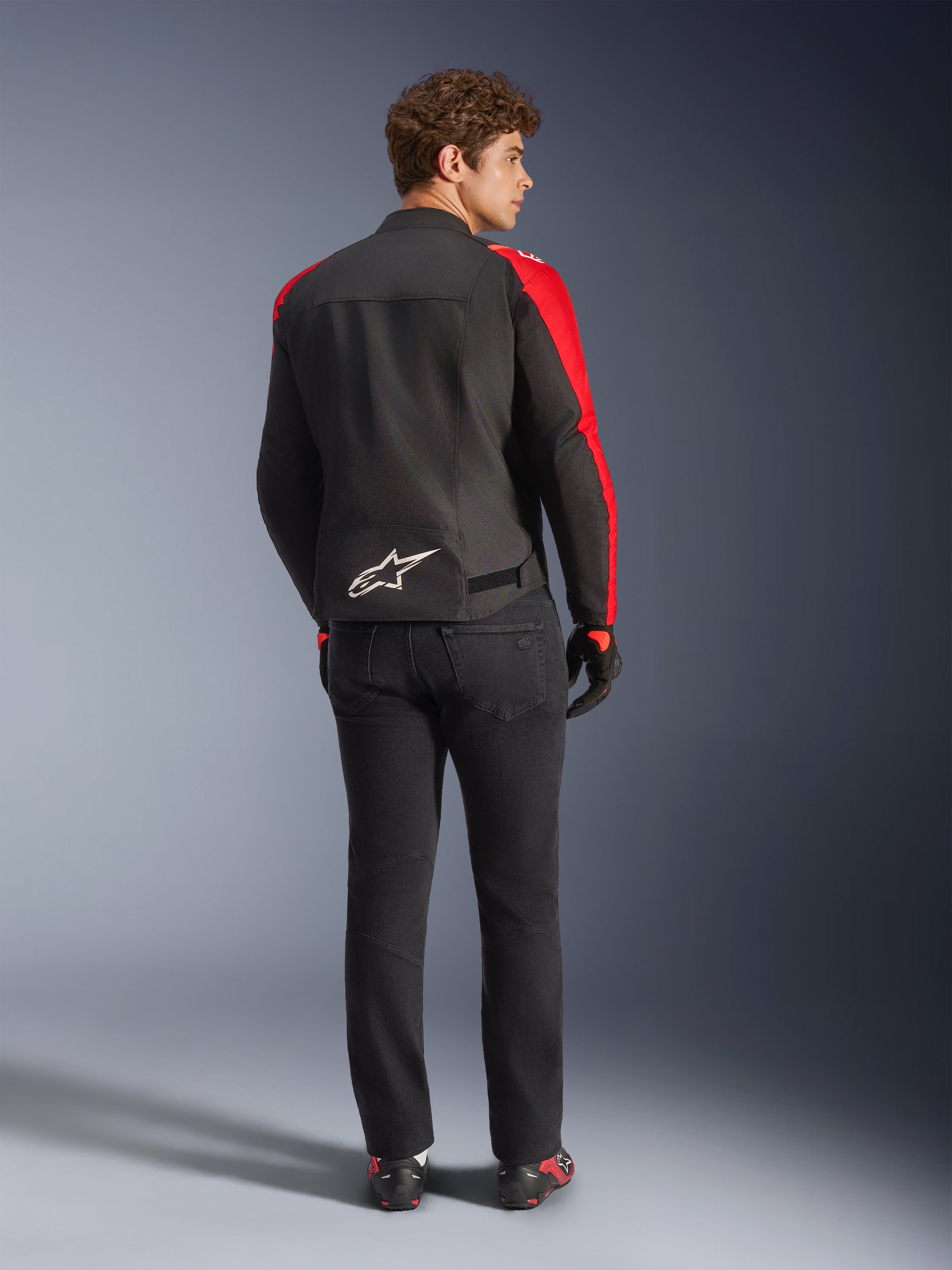 Veste imperméable Alpinestars T-SPS V2, noir, rouge et rouge fluo, vue de dos sur un modèle masculin, logo Alpinestars blanc en bas du dos, portée avec un pantalon de moto noir et des gants, veste de moto en textile imperméable au style sportif