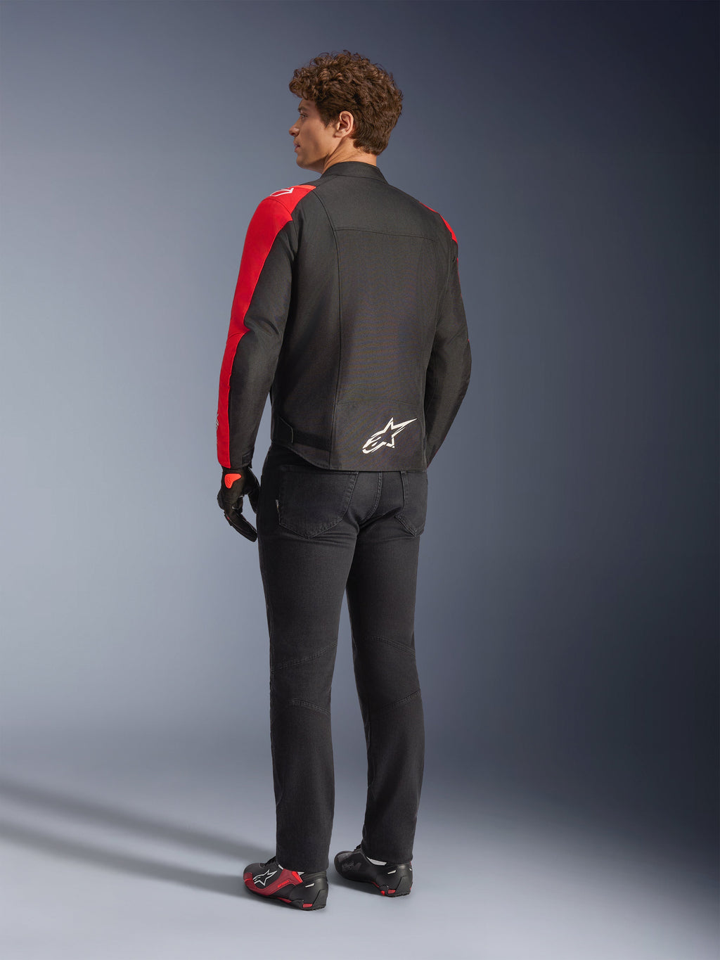 Veste imperméable Alpinestars T-SPS V2 en noir et rouge fluo, vue de dos sur un modèle, portée avec un pantalon de moto noir et des chaussures de pilotage, avec un logo Alpinestars blanc en bas du dos, design textile à coupe sport