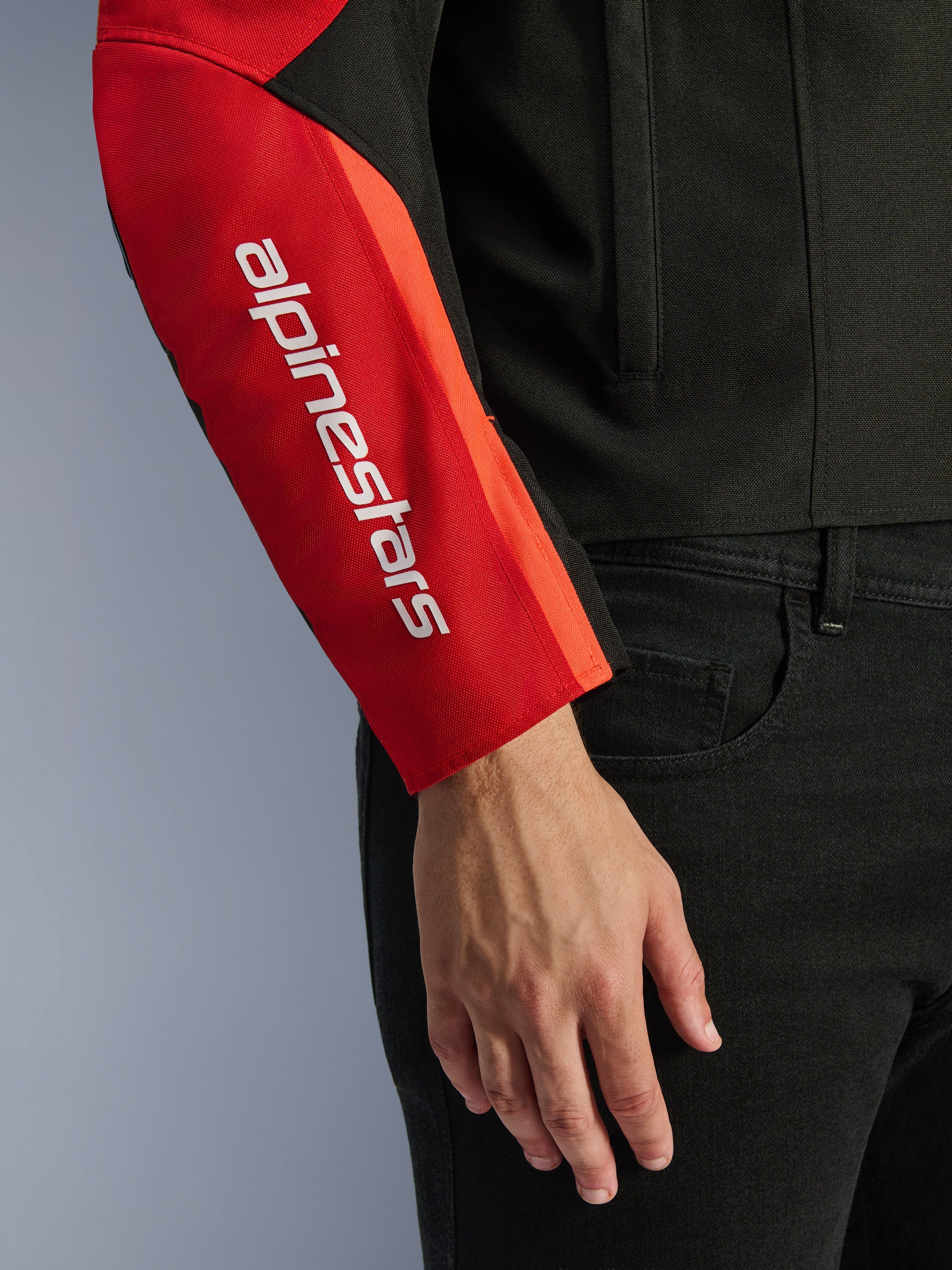 Veste imperméable Alpinestars T-SPS V2, veste de moto, noir et rouge, gros plan de la manche présentant un logo de marque blanc vertical sur un empiècement rouge, construction en textile durable avec un design inspiré du sport.