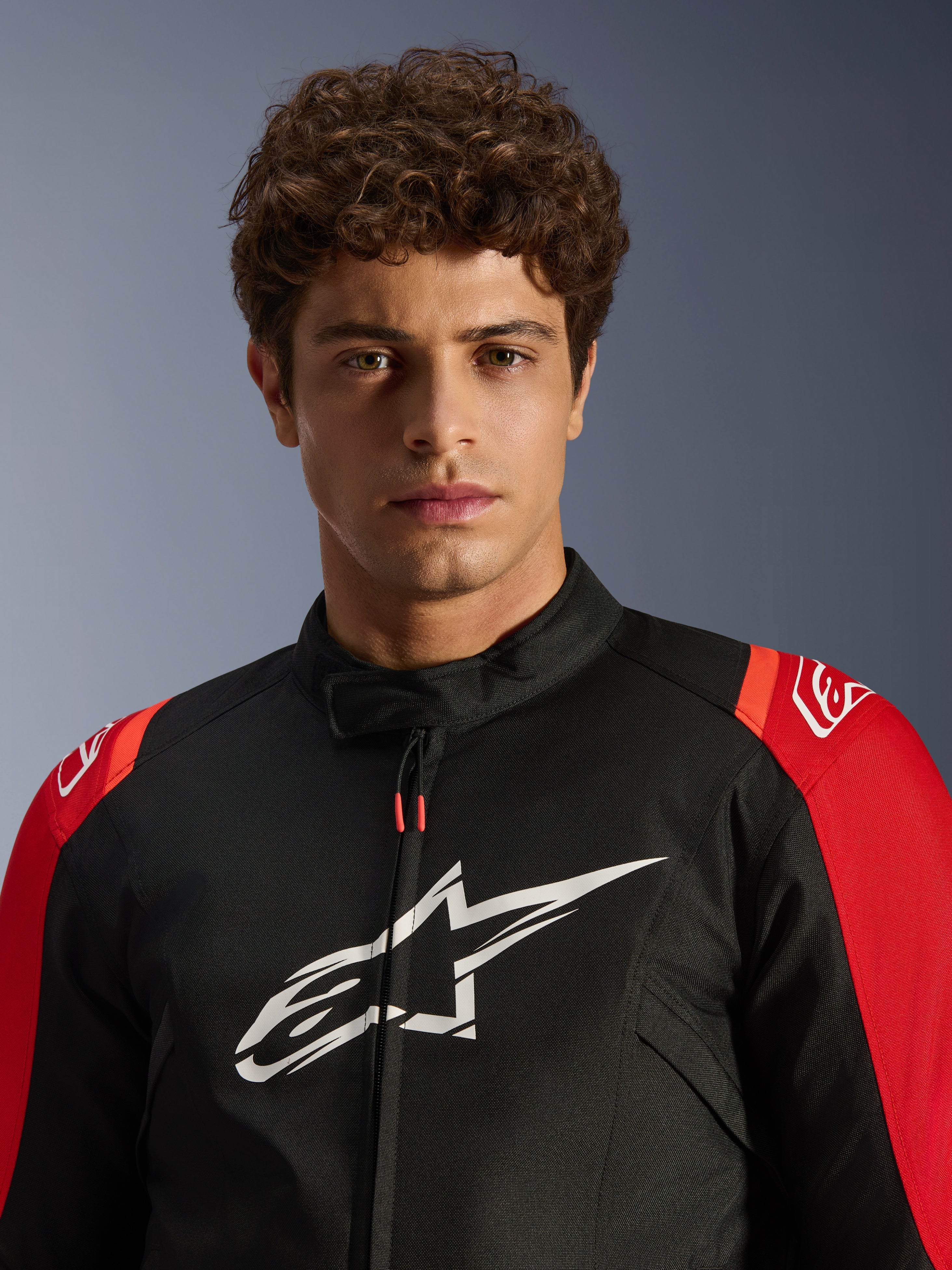 Modèle masculin portant la veste imperméable Alpinestars T-SPS V2, noire avec accents rouges et logo blanc, portrait en gros plan, veste de moto de sport en textile