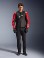 Modèle masculin portant la veste imperméable Alpinestars T-SPS V2, noire avec des manches rouges et rouge fluo, logo blanc sur la poitrine, associée à un pantalon noir, des gants et des chaussures de moto noirs, design sportif d'équipement de route pour moto