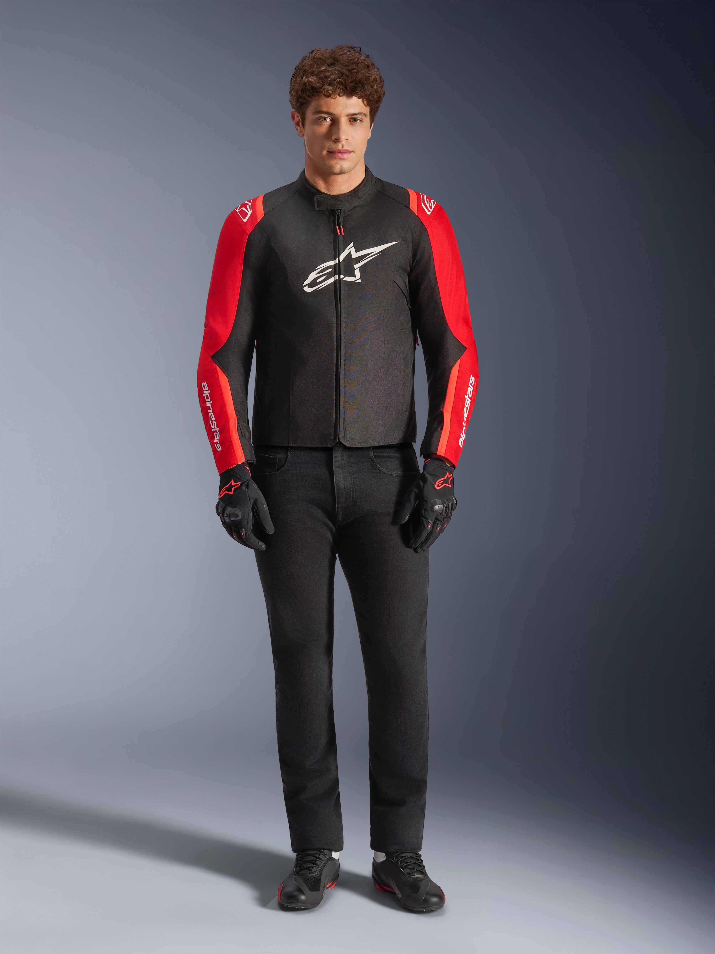 Modèle masculin portant la veste imperméable Alpinestars T-SPS V2, noire avec des manches rouges et rouge fluo, logo blanc sur la poitrine, associée à un pantalon noir, des gants et des chaussures de moto noirs, design sportif d'équipement de route pour moto