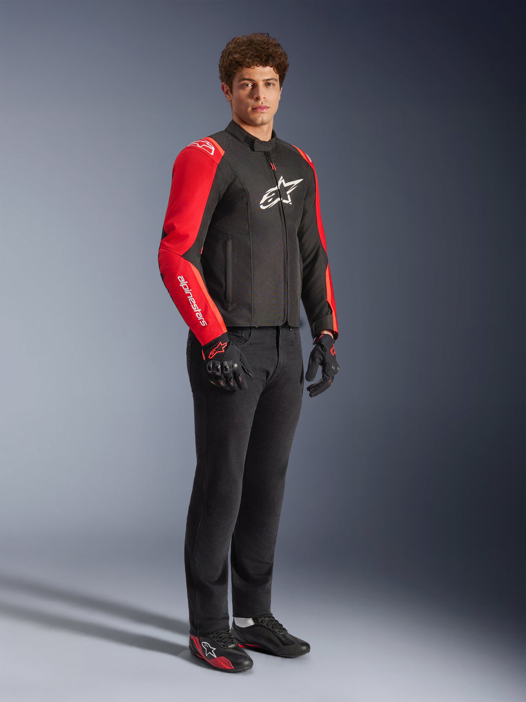Mannequin homme portant la veste imperméable Alpinestars T-SPS V2 en Noir et Rouge Fluo, dotée de manches rouges avec marquage blanc et d'un grand logo blanc sur la poitrine, associée à un pantalon de moto noir, des gants et des chaussures de moto à profil bas, vue studio en pied.