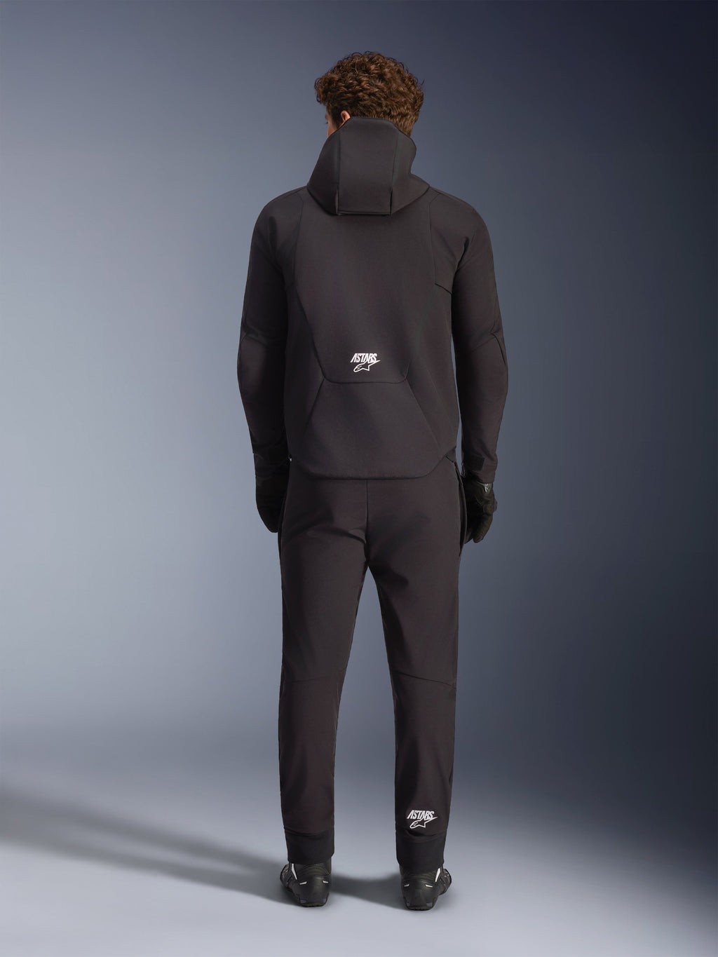 Veste imperméable Alpinestars Aeroshell, noire, vue de dos sur un mannequin portant un pantalon de moto et des gants noirs assortis, design softshell sport à capuche avec logos ASTARS blancs, vêtement de moto élégant et moderne
