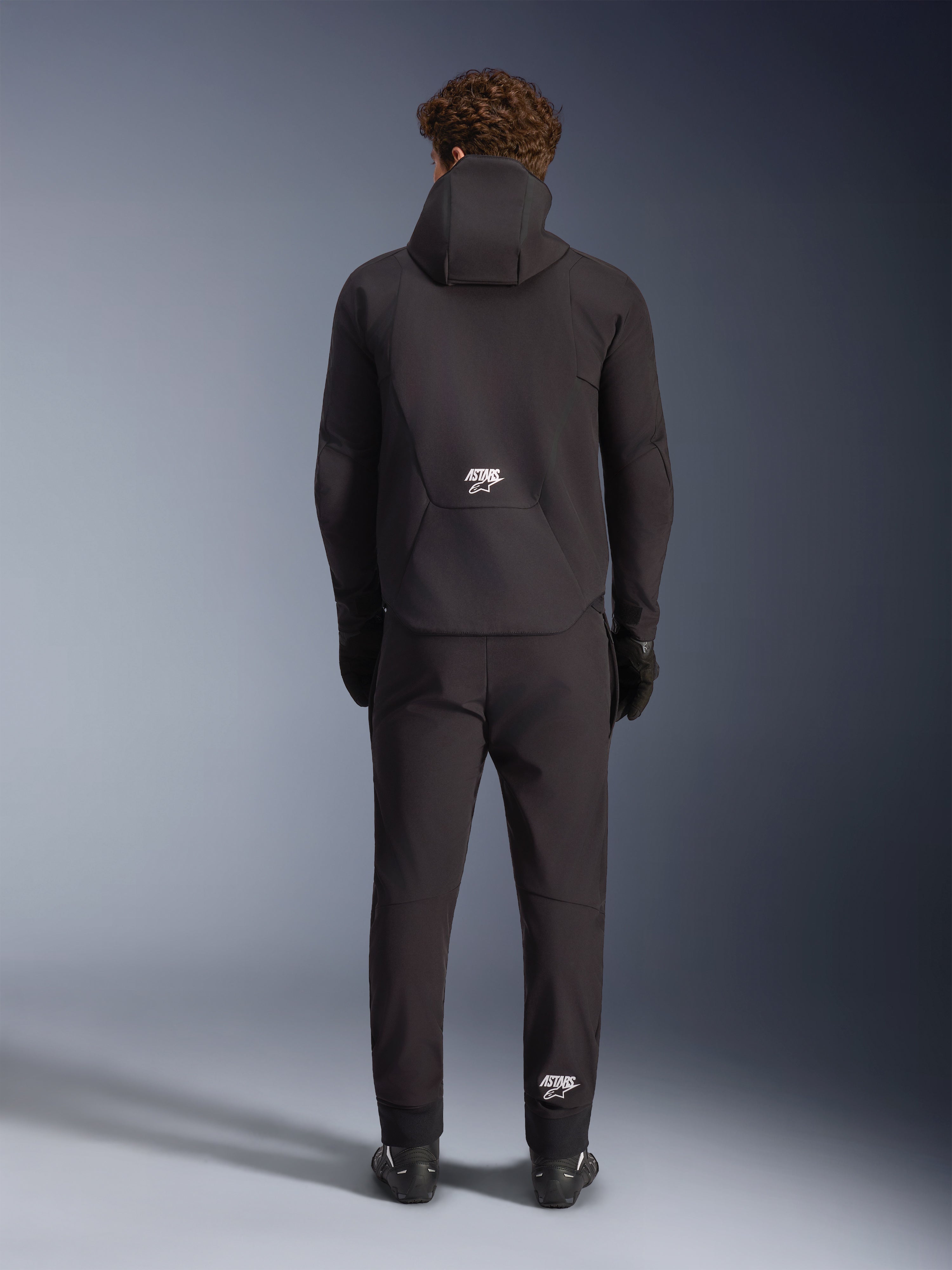 Veste imperméable Alpinestars Aeroshell, noire, vue de dos sur un mannequin portant un pantalon de moto et des gants noirs assortis, design softshell sport à capuche avec logos ASTARS blancs, vêtement de moto élégant et moderne
