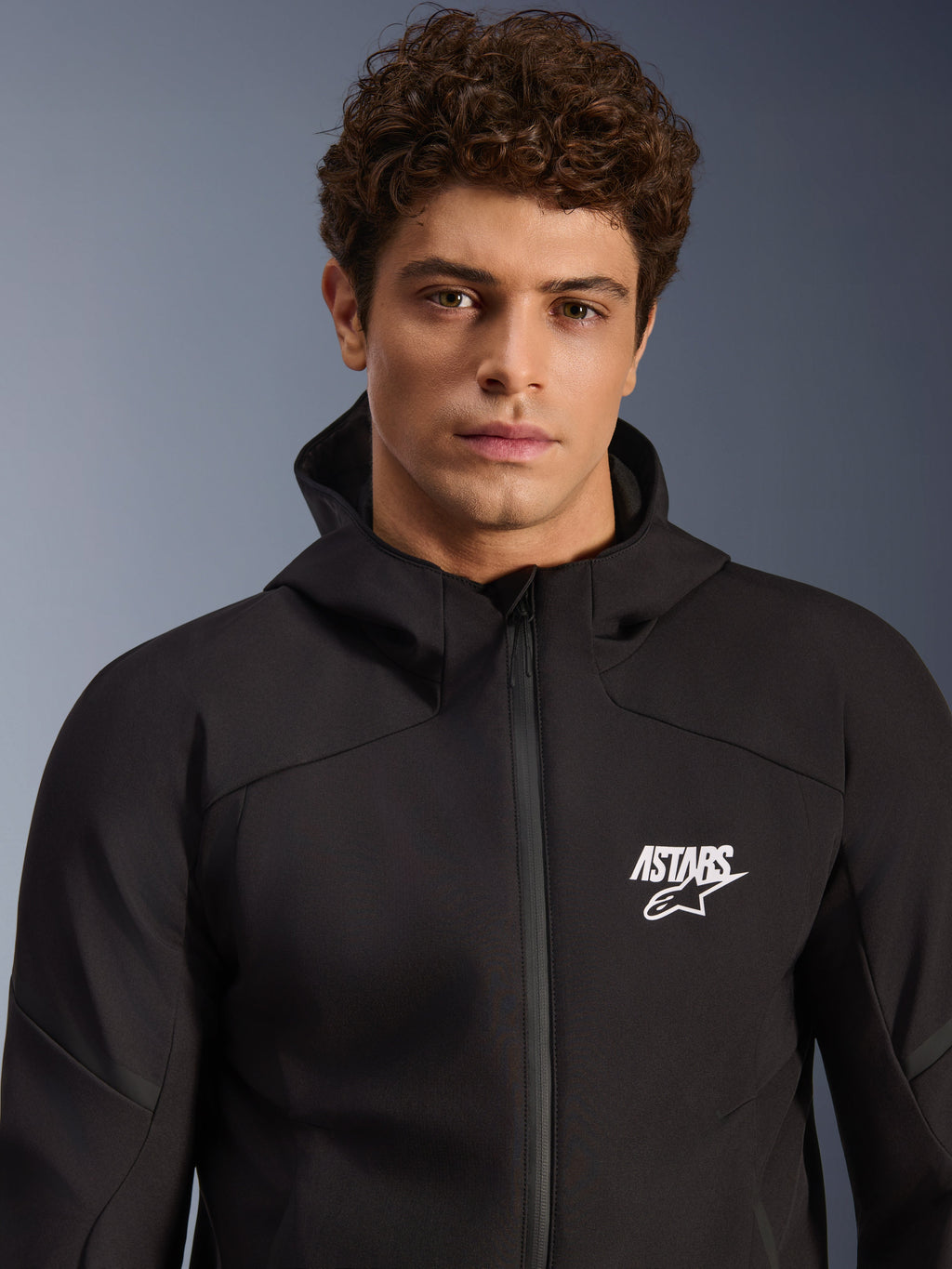 Modèle masculin portant la veste imperméable Alpinestars Aeroshell WP, veste softshell noire en textile avec capuche, logo ASTARS blanc sur la poitrine, fermeture éclair frontale imperméable, portrait moderne en studio pour équipement de route moto