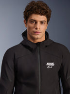 Modèle masculin portant la veste imperméable Alpinestars Aeroshell WP, veste softshell noire en textile avec capuche, logo ASTARS blanc sur la poitrine, fermeture éclair frontale imperméable, portrait moderne en studio pour équipement de route moto