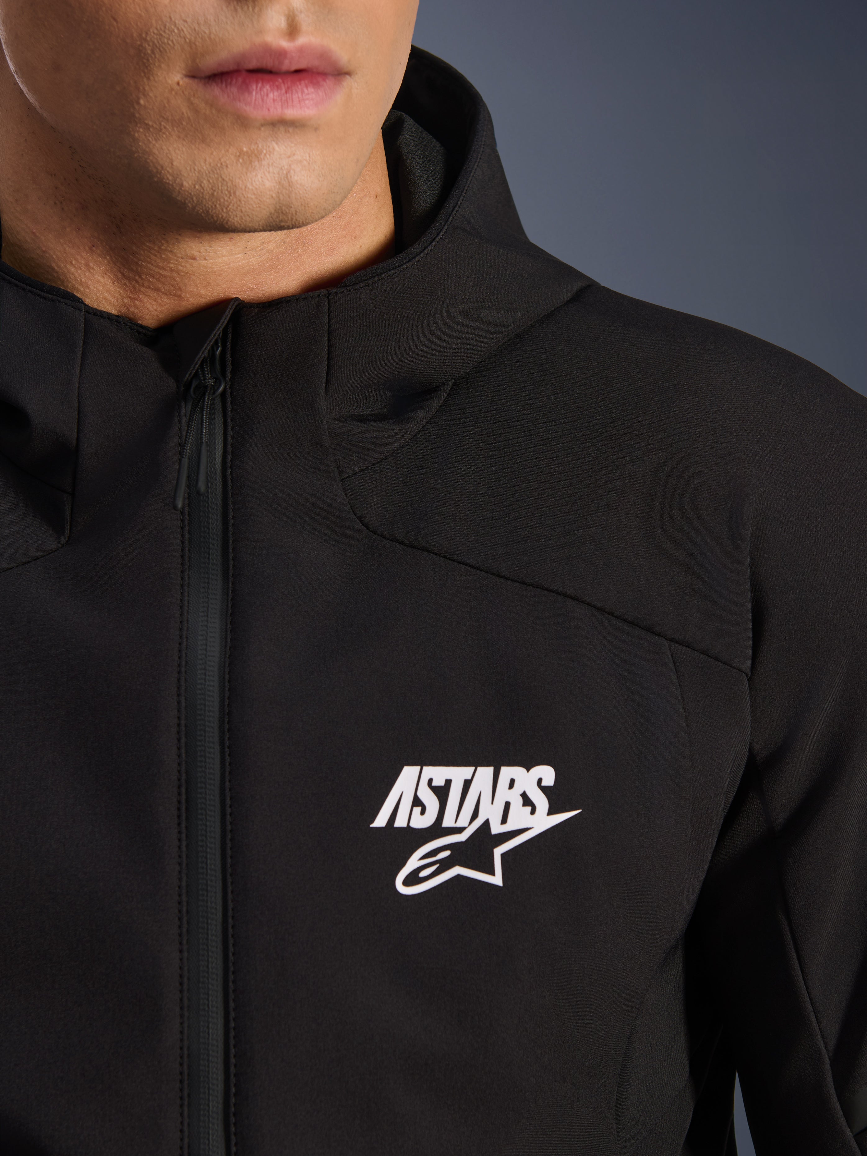 Veste Alpinestars Aeroshell Waterproof, veste de moto softshell noire, vue rapprochée de la poitrine avec logo étoile ASTARS blanc et fermeture éclair avant imperméable, capuche intégrée, design sportif moderne