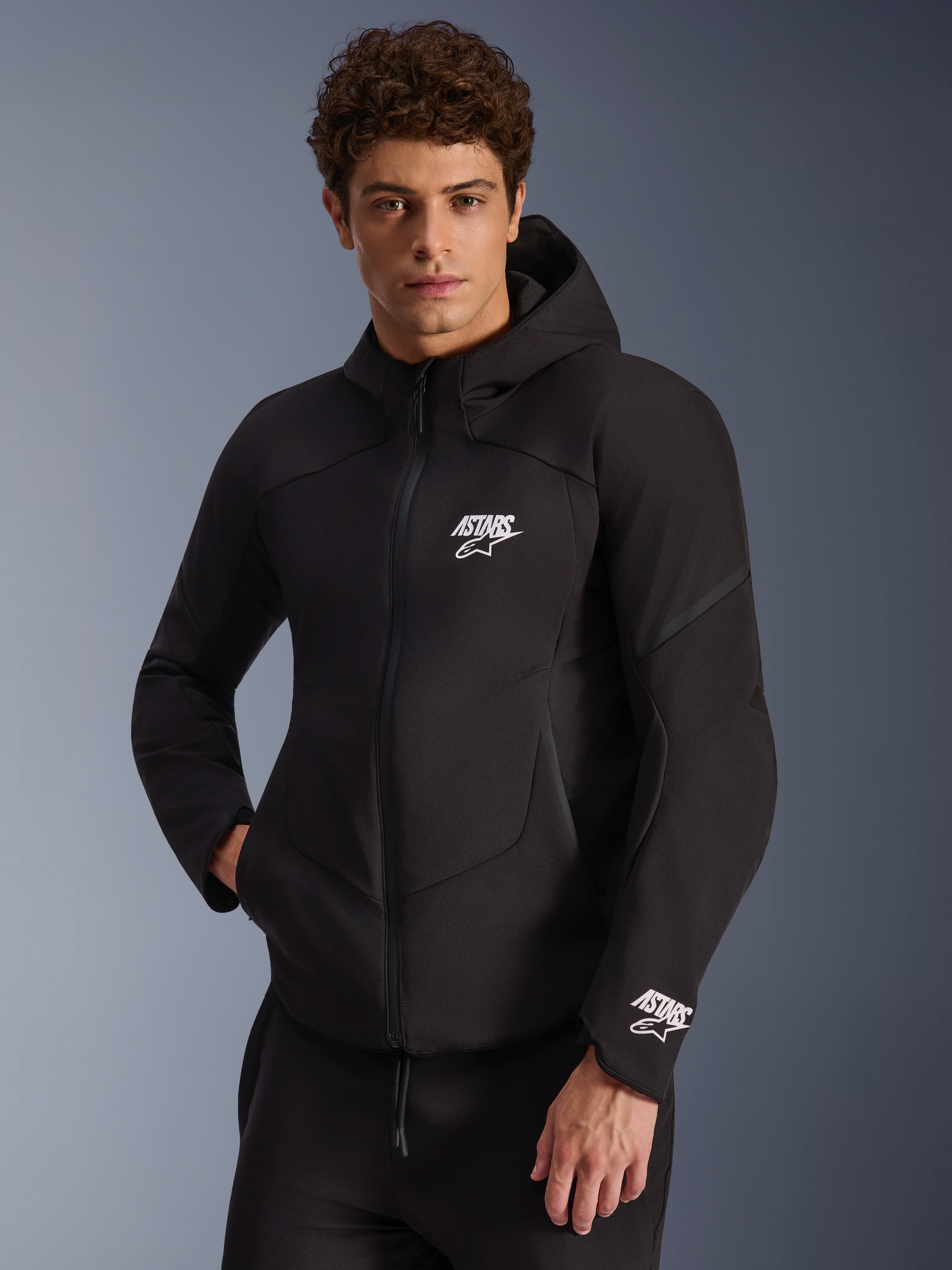 Veste imperméable Alpinestars Aeroshell, sweat à capuche de moto en softshell noir, logo ASTARS blanc sur la poitrine et le poignet, design épuré avec capuche intégrée et fermeture éclair imperméable, équipement moderne d'inspiration sportive pour la conduite urbaine