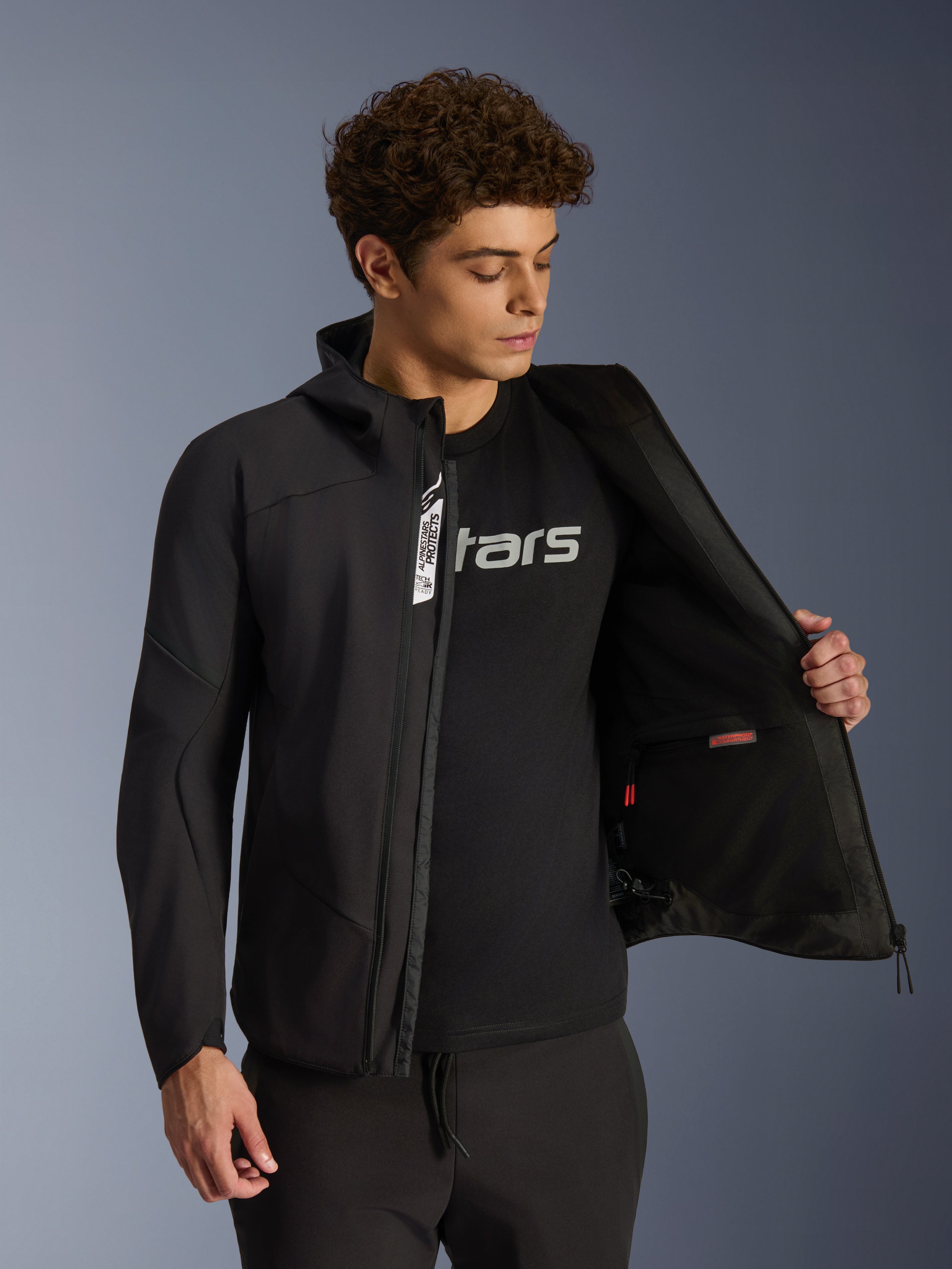 Veste Alpinestars Aeroshell Waterproof, veste de moto softshell noire, dézippée laissant apparaître la poche intérieure et la doublure, portée par un mannequin avec un t-shirt noir à logo, design à capuche pour un style urbain et décontracté.