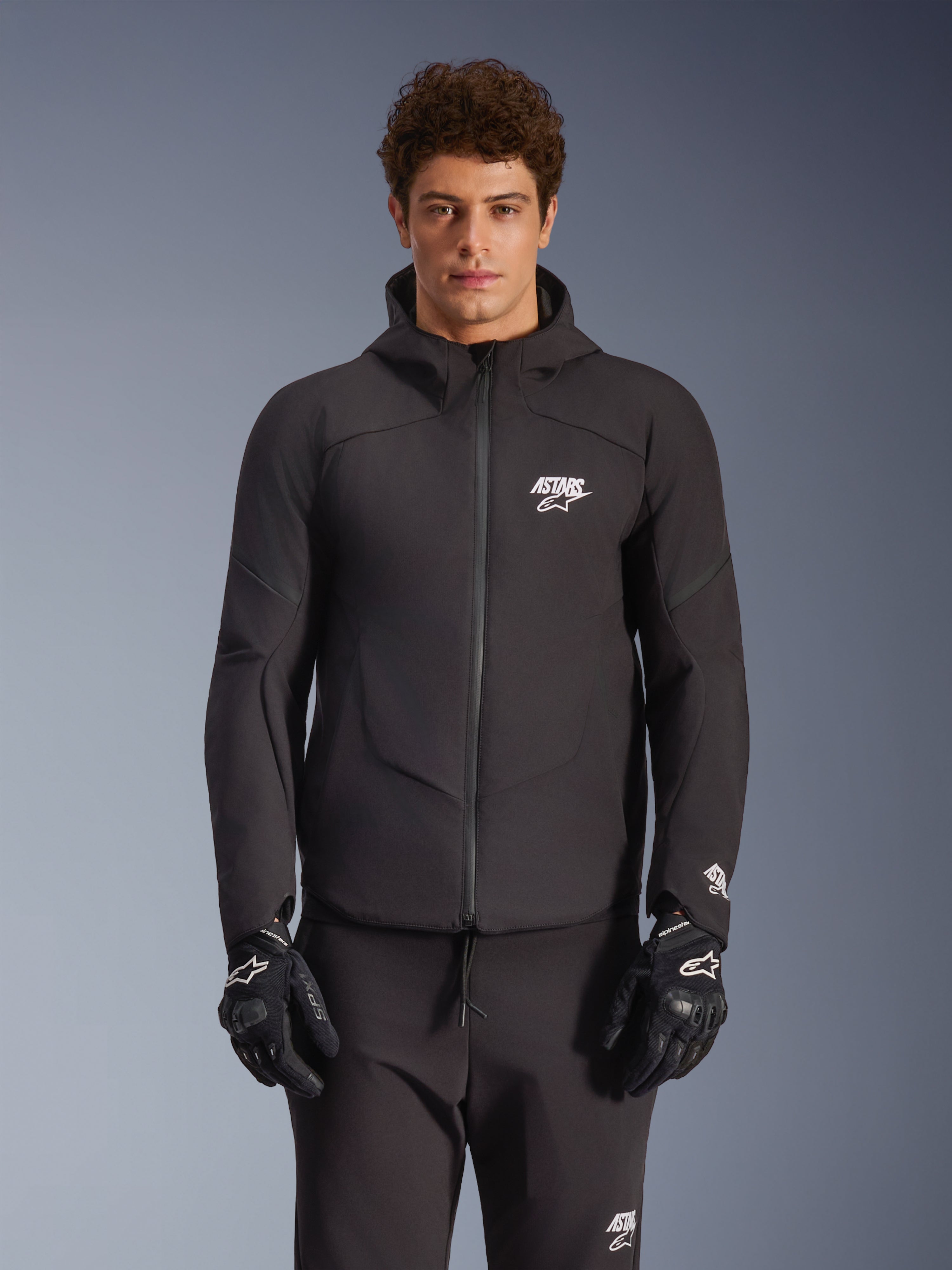 Veste imperméable Alpinestars Aeroshell, noire, veste de moto urbaine, design à capuche avec fermeture éclair frontale, logos blancs sur la poitrine et les manches, portée avec un pantalon et des gants noirs assortis