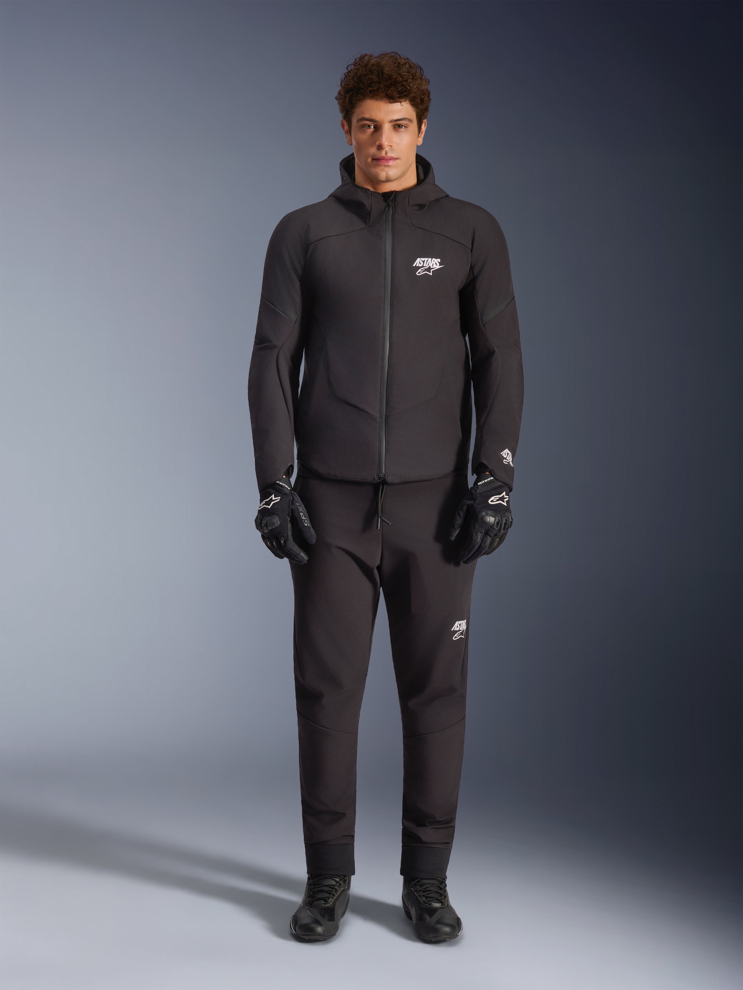 Mannequin homme portant la veste imperméable Alpinestars Aeroshell WP en noir, avec pantalon technique et gants de moto noirs assortis, design softshell à capuche avec détails de logo blancs, tenue moderne inspirée du sport moto pour la conduite sur route