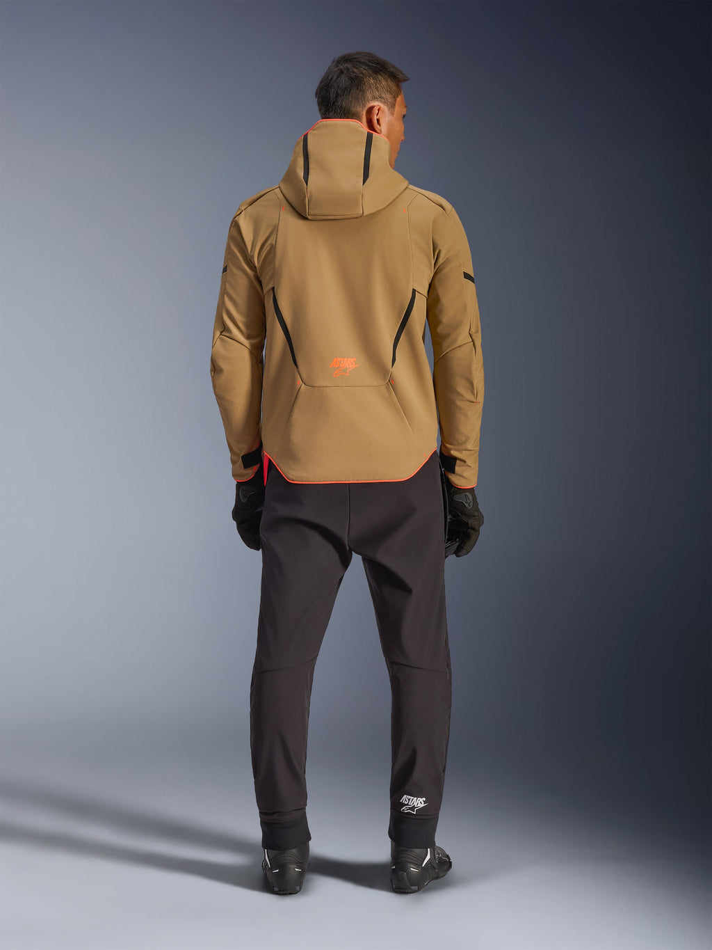 Veste Alpinestars Aeroshell Waterproof en Kaki Foncé Noir Orange Fluo, vue de dos d'un homme portant la veste softshell à capuche, avec liserés noirs et logo orange, associée à un pantalon technique noir et des gants.