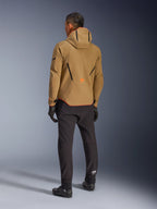 Veste imperméable Alpinestars Aeroshell, Dark Khaki Black Orange Fluo, vue arrière d'un motard portant la veste softshell à capuche, associée à un pantalon et des gants noirs, présentant des accents géométriques noirs et un logo orange en bas du dos.