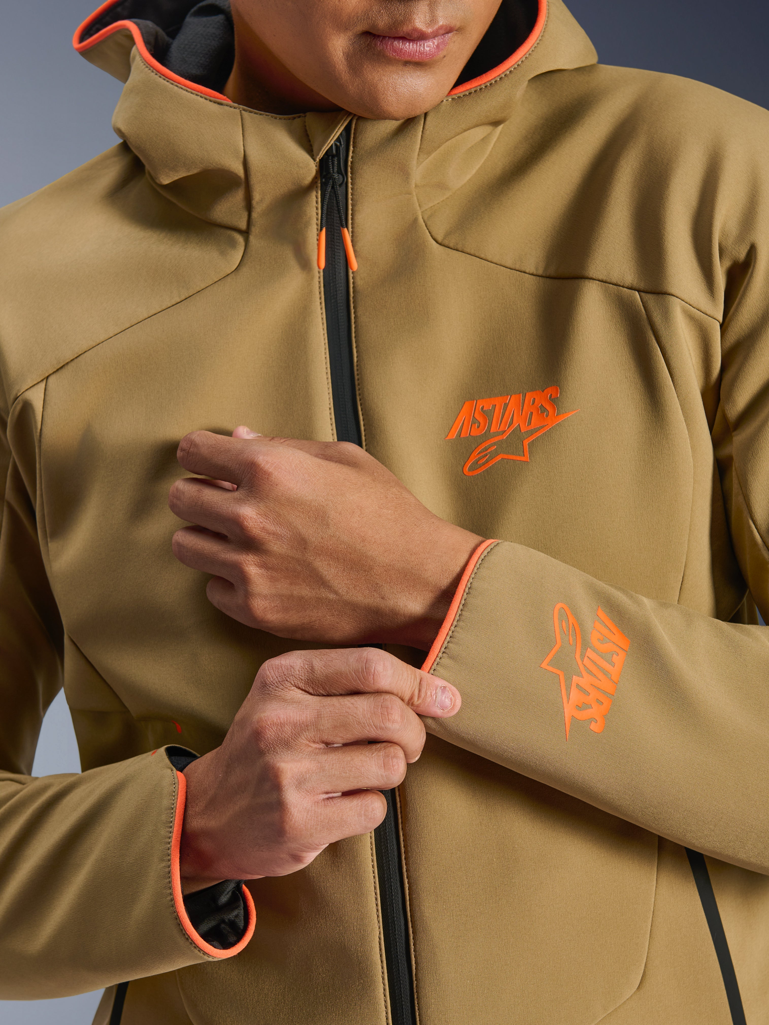 Veste imperméable Alpinestars Aeroshell, kaki foncé et orange fluo, gros plan sur l'avant et la manche présentant des logos ASTARS orange, des liserés orange sur la capuche et les poignets, et une fermeture éclair imperméable noire.