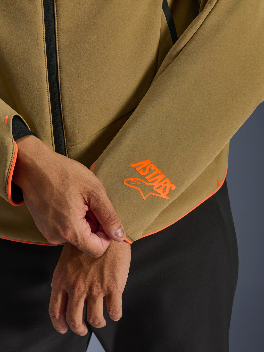 Veste imperméable Alpinestars Aeroshell, kaki foncé avec accents orange fluo, gros plan sur le poignet de la manche et l'ourlet inférieur avec le logo Astars orange vif, liseré orange fluo et détail de la fermeture éclair avant imperméable, construction softshell sport