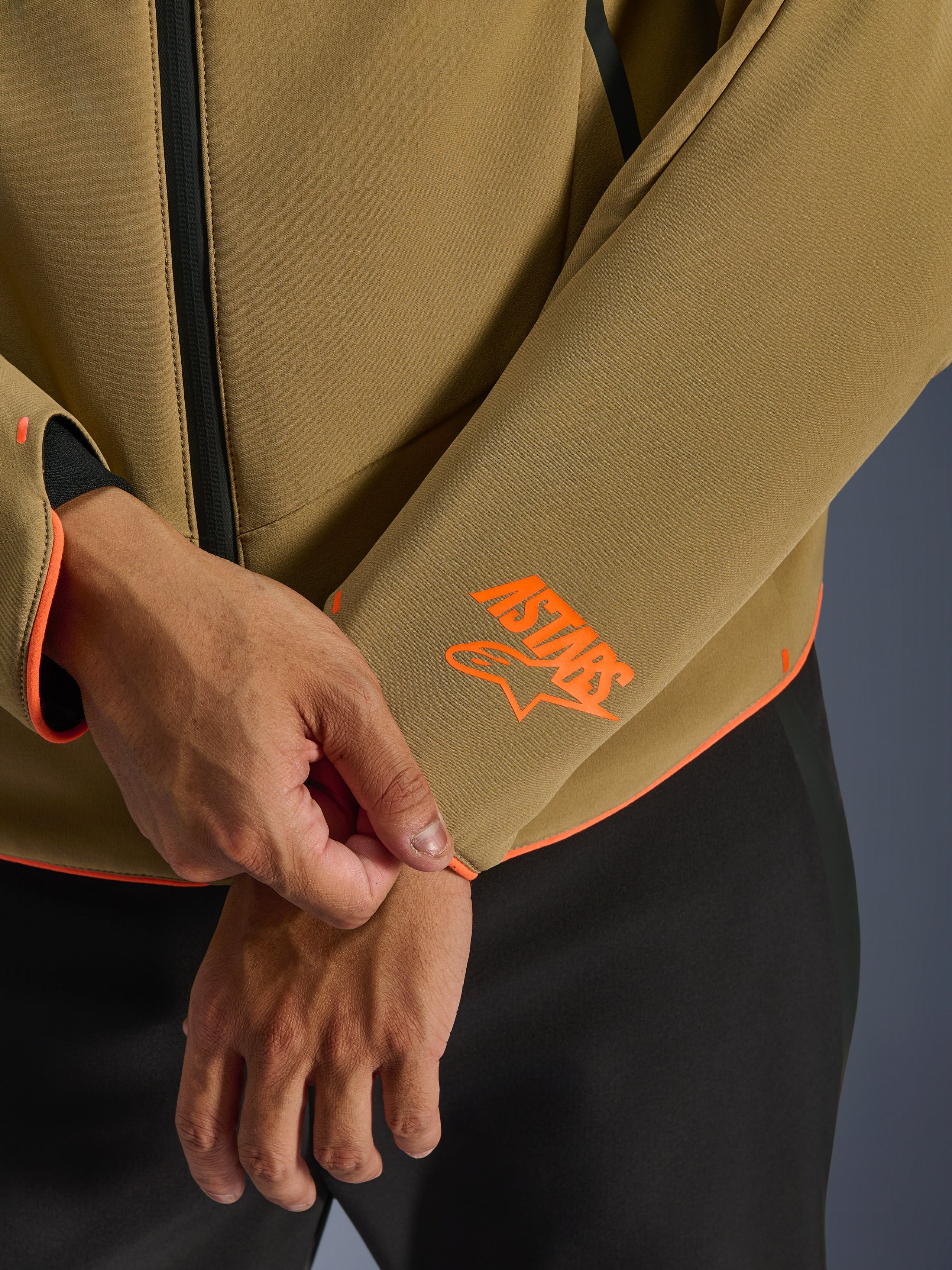 Veste imperméable Alpinestars Aeroshell, kaki foncé avec accents orange fluo, gros plan sur le poignet de la manche et l'ourlet inférieur avec le logo Astars orange vif, liseré orange fluo et détail de la fermeture éclair avant imperméable, construction softshell sport