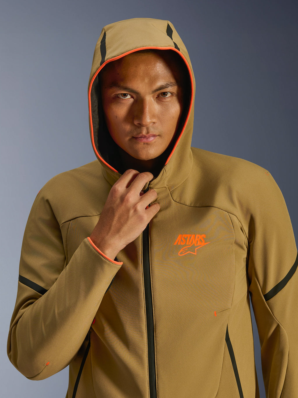 Veste Alpinestars Aeroshell WP, kaki foncé, noir et orange fluo, mannequin portant la veste avec la capuche relevée, avec bordure orange fluo et logo ASTARS orange, accents noirs, softshell imperméable au style sportif moderne
