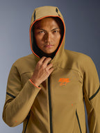 Veste Alpinestars Aeroshell WP, kaki foncé, noir et orange fluo, mannequin portant la veste avec la capuche relevée, avec bordure orange fluo et logo ASTARS orange, accents noirs, softshell imperméable au style sportif moderne