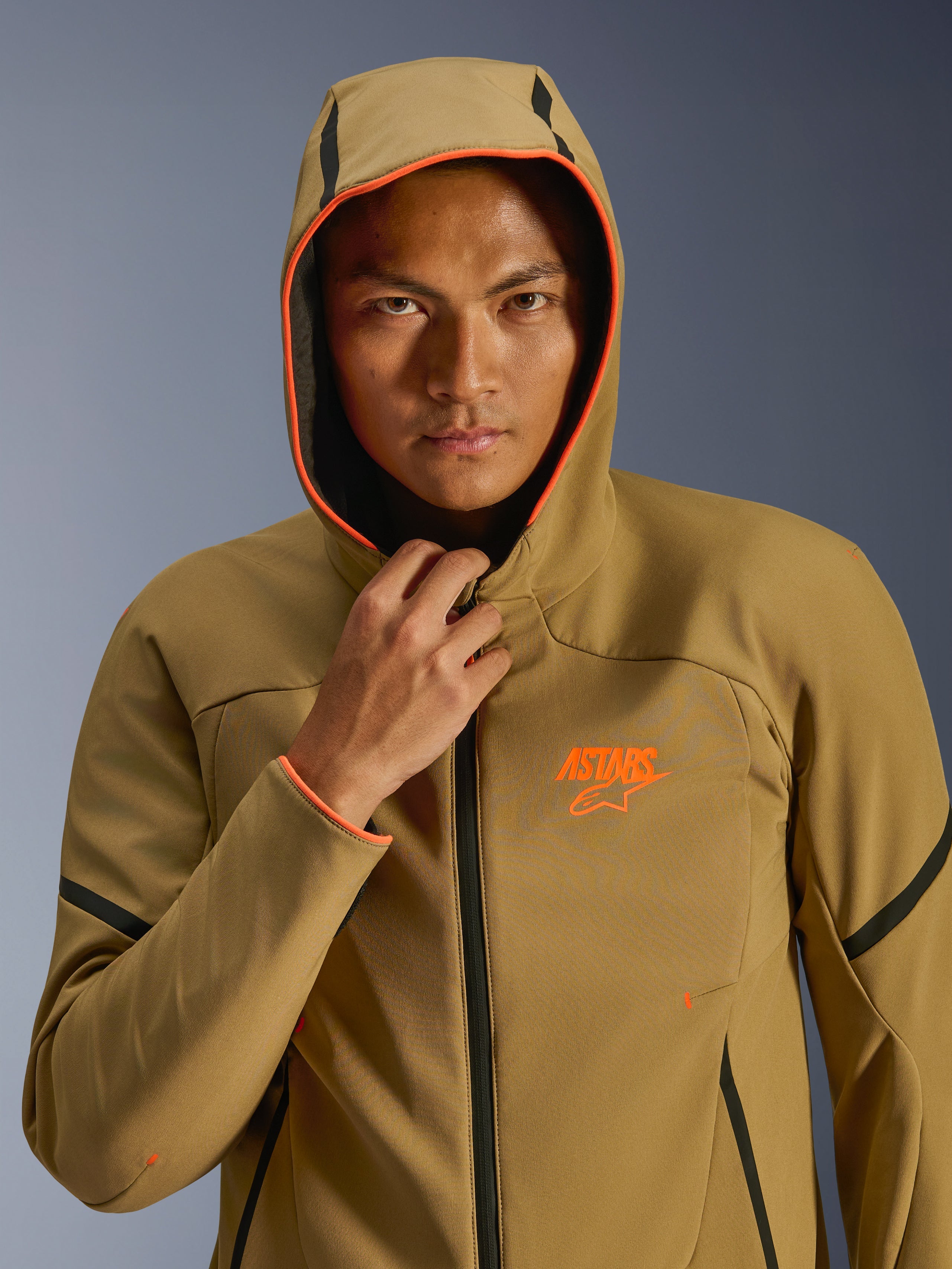 Veste Alpinestars Aeroshell WP, kaki foncé, noir et orange fluo, mannequin portant la veste avec la capuche relevée, avec bordure orange fluo et logo ASTARS orange, accents noirs, softshell imperméable au style sportif moderne