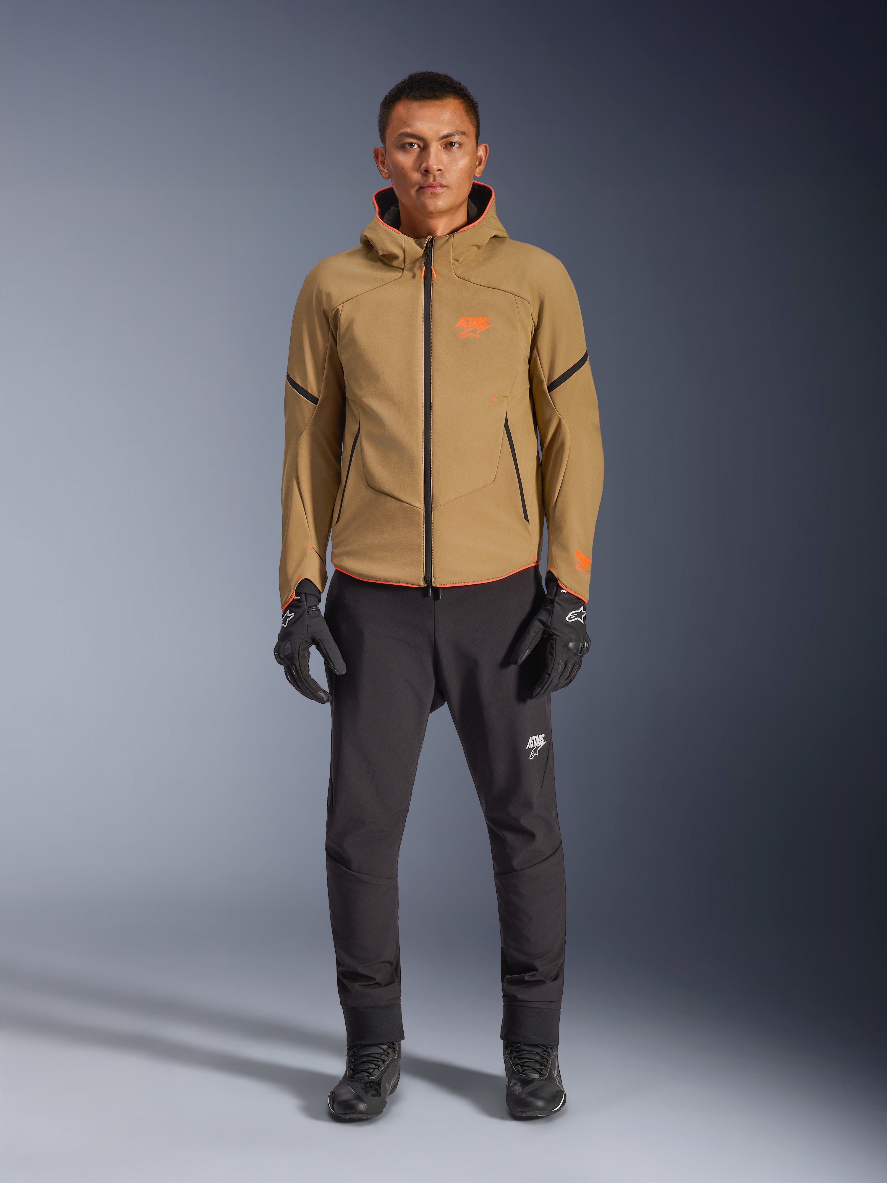 Mannequin homme portant la veste imperméable Alpinestars Aeroshell WP en Kaki Foncé avec accents Noir et Orange Fluo, logo orange sur la poitrine et design à capuche, associée à un pantalon noir et des gants de moto noirs, style sport-touring décontracté