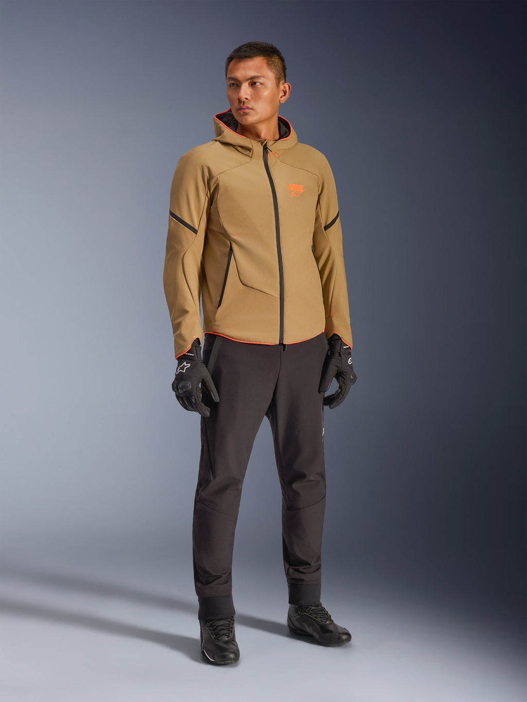 Veste imperméable Alpinestars Aeroshell en coloris kaki foncé, noir et orange fluo, portée par un mannequin avec un équipement assorti, dotée d'un design softshell à capuche, de fermetures éclair étanches et de logos orange vif.