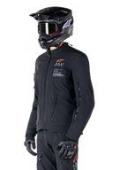 Veste Alpinestars AMT-8 Stretch Drystar® XF, noire, portée par un pilote avec pantalon d'aventure et gants assortis, comprenant un casque de motocross noir et des lunettes, ouvertures de ventilation zippées sur la poitrine et les bras, marquage Alpinestars Motion Technology avec logo orange, équipement d'aventure-touring haut de gamme