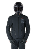 Veste Alpinestars AMT-8 Stretch Drystar® XF, veste de moto noire, avec logo Alpinestars orange et marquage AMT gris, col haut avec fermeture éclair frontale et ventilations, portée avec un casque et des gants noirs, équipement de protection pour le touring d'aventure