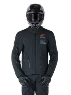 Veste Alpinestars AMT-8 Stretch Drystar® XF, veste de moto noire, avec logo Alpinestars orange et marquage AMT gris, col haut avec fermeture éclair frontale et ventilations, portée avec un casque et des gants noirs, équipement de protection pour le touring d'aventure