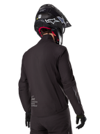 Doublure Alpinestars AMT-8 Stretch Drystar® XF, doublure de veste imperméable noire, vue arrière d'un pilote portant un casque noir et des gants, avec le marquage Alpinestars Motion Technology sur le bas du dos, design minimaliste et fonctionnel pour la conduite aventure.