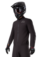 Pilote portant la doublure Alpinestars AMT-8 Stretch Drystar® XF, veste stretch noire coupe-vent et imperméable, arborant le logo AMT sur la poitrine et le marquage Drystar XF sur la manche, associée à un casque d'aventure noir et des gants de protection.