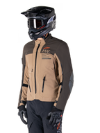 Veste Alpinestars AMT-8 Stretch Drystar® XF, veste de moto d'aventure, brun foncé et kaki foncé, portée avec un casque tout-terrain noir et des lunettes, arborant le marquage Alpinestars Motion Technology et des ouvertures de ventilation zippées.