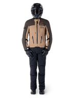 Veste Alpinestars AMT-8 Stretch Drystar® XF, veste de moto adventure, brun foncé et kaki foncé, portée avec un casque noir, un masque, des gants noirs, un pantalon noir et des bottes noires, dotée d'orifices de ventilation sur la poitrine et du marquage AMT, équipement coordonné pour le touring d'aventure