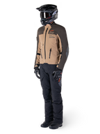 Pilote d'aventure portant un équipement complet, veste Alpinestars AMT-8 Stretch Drystar® XF en brun foncé et kaki foncé, associée à un pantalon d'aventure noir, un casque, des gants et des bottes noirs, avec logo AMT orange et fermetures éclair de ventilation techniques pour le touring longue distance.
