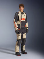 Male model wearing the Alpinestars Halo Pro Drystar® XF Laminated Jacket in Mastic Black, featuring beige and black panels with red accents, paired with matching adventure pants and black gloves, designed for all-terrain adventure motorcycle riding.